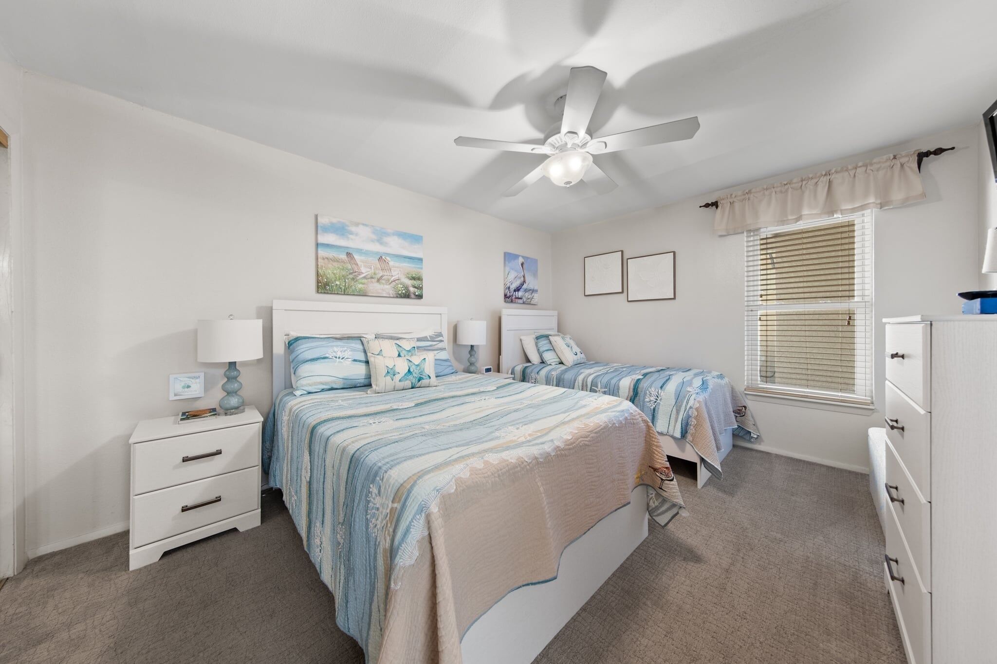 A bedroom with two beds, a ceiling fan, a nightstand, a dresser, and a window with blinds.
