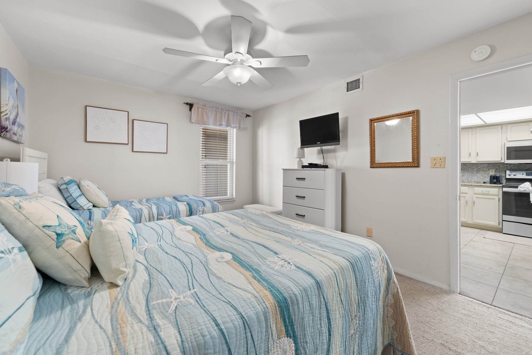 A bedroom with a bed, a ceiling fan, a TV, a mirror, and a view into the kitchen.