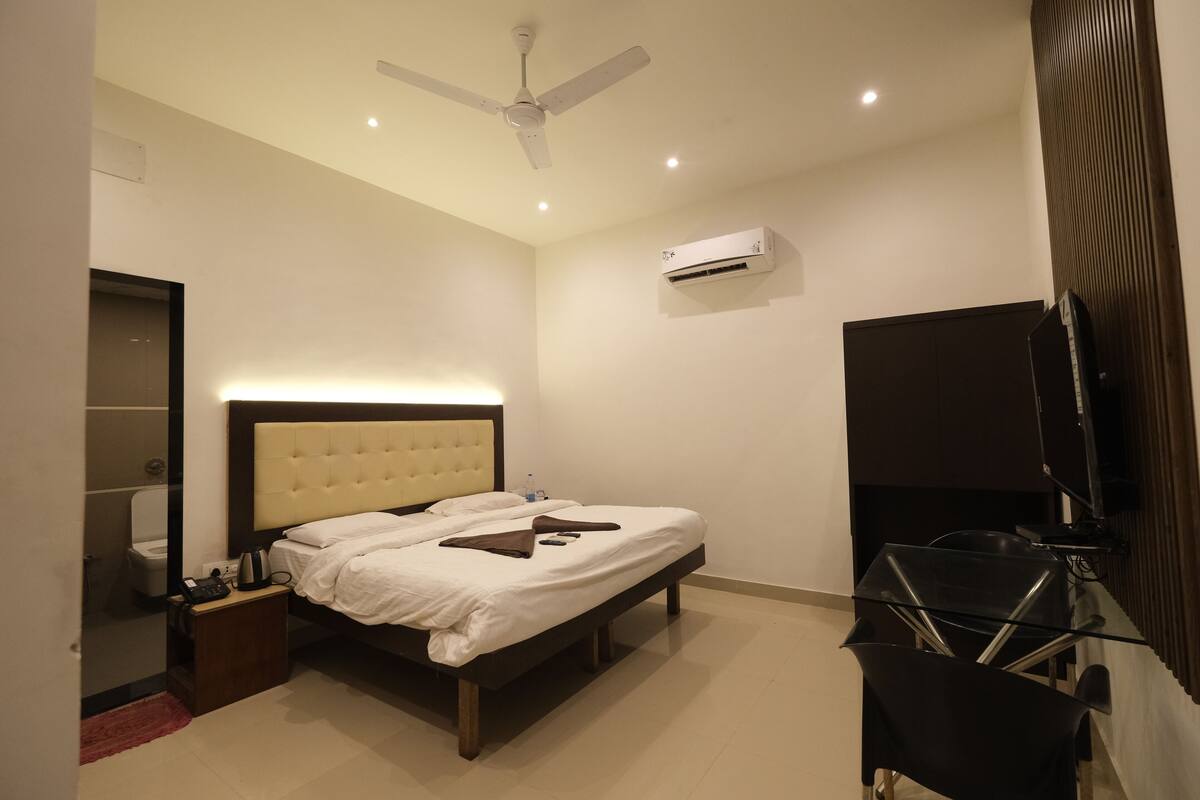 A bedroom with a bed, a ceiling fan, an air conditioning unit, a television, and a small table with a kettle.