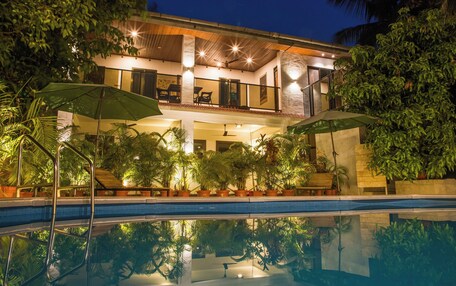 A two-story house with a swimming pool, surrounded by lush greenery and potted plants.