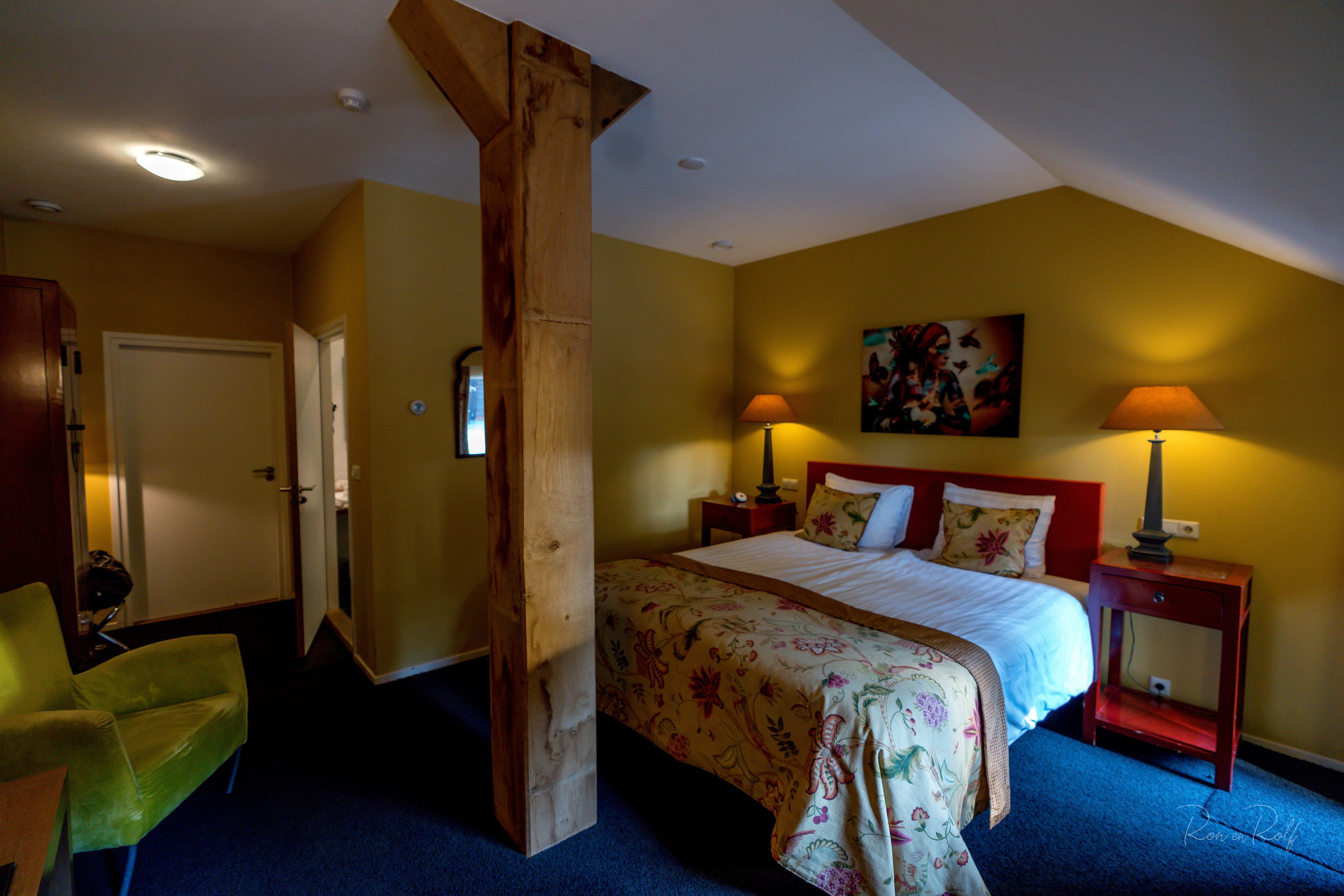 A bedroom with a bed, two bedside tables with lamps, a chair, a wooden pillar, and a door.