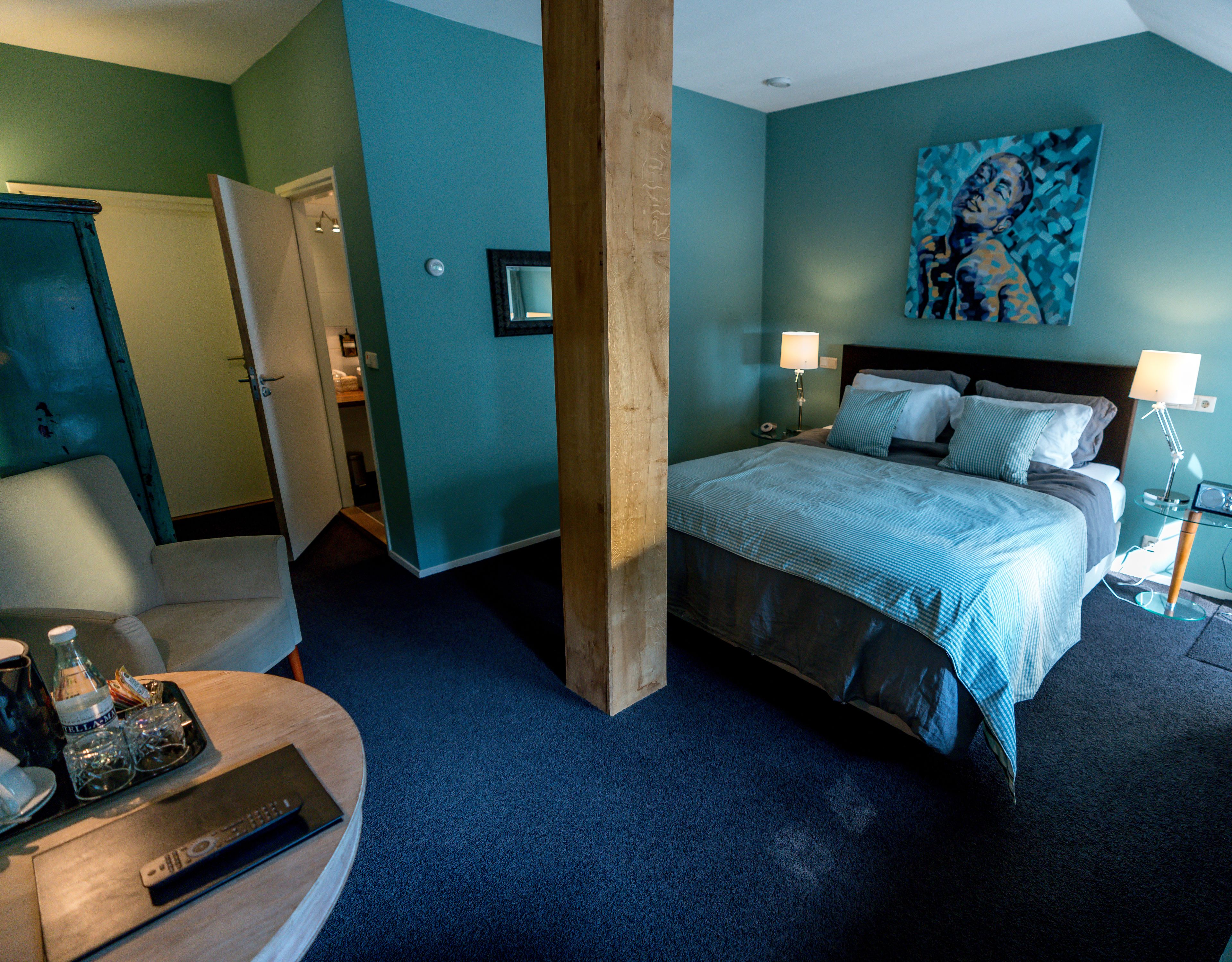 A hotel room with a large bed, a chair, a desk with a remote control, and a painting on the wall.