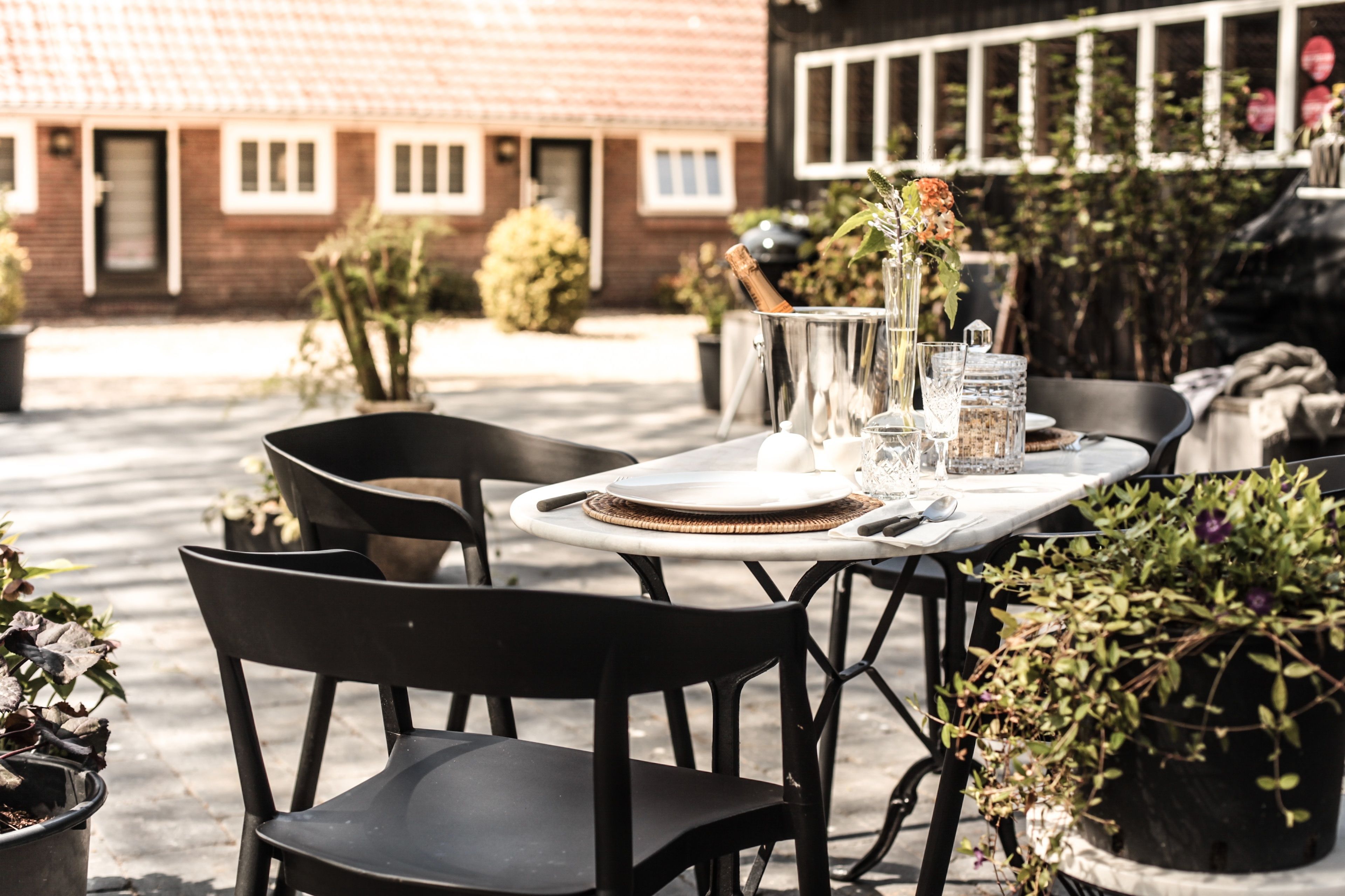 An outdoor dining area with a table set for two, surrounded by potted plants and a brick building in the background.