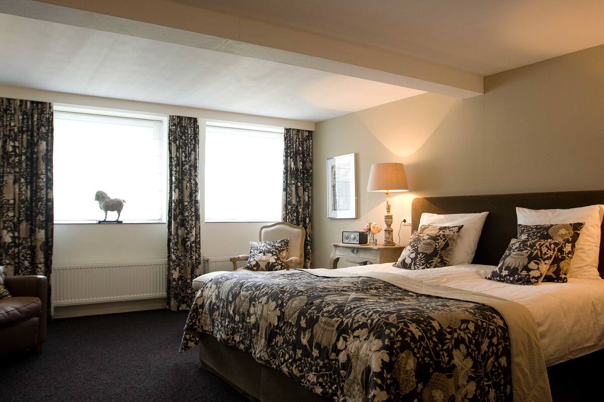 Une chambre d’hôtel avec un grand lit, une chaise, une table de chevet avec une lampe et une fenêtre avec des rideaux.