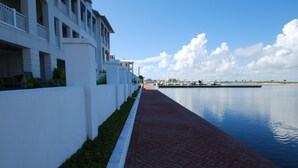 Un paseo marítimo con sendero de ladrillo rojo, edificios blancos con balcones y un puerto deportivo al fondo.