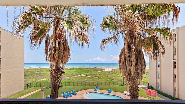 A pool area with lounge chairs and palm trees, overlooking a beach and ocean. - Beach House Unit #302 2 Bedroom Condo by RedAwning (South Padre Island)