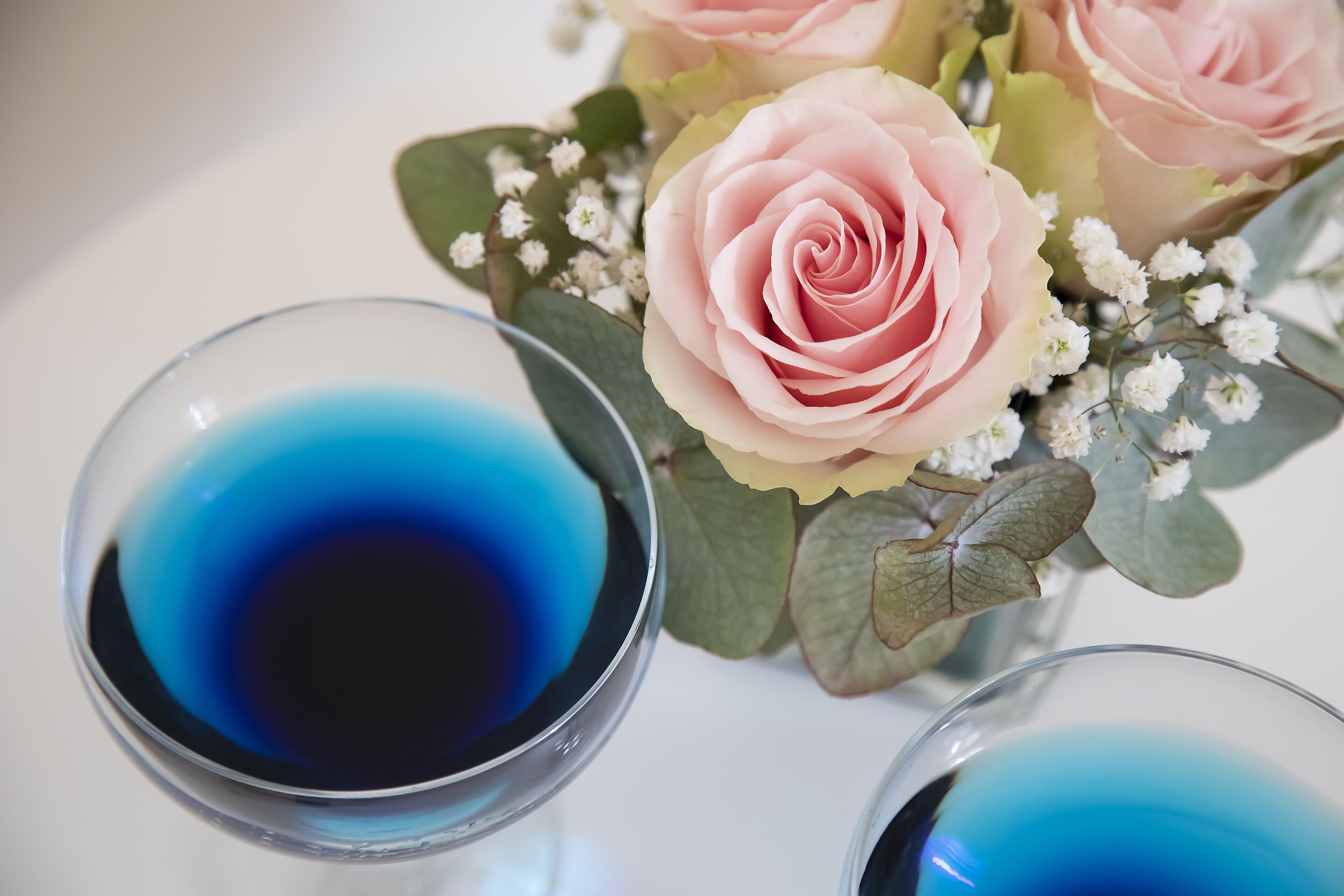 Zwei Gläser mit blauer Flüssigkeit, eine rosarote Rose und weiße Blumen.