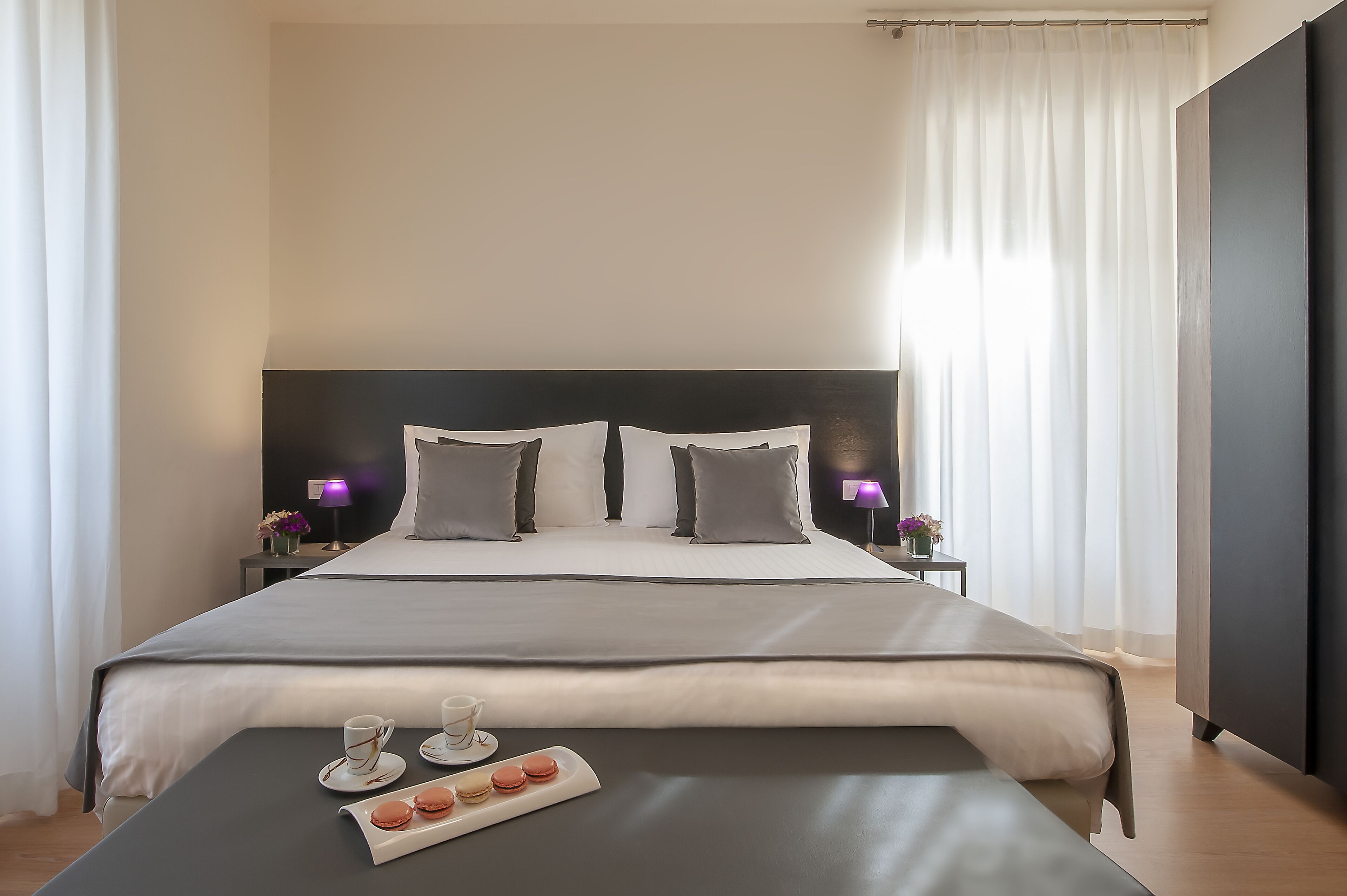 Una habitación de hotel moderna con una cama grande, lámparas de noche, una mesita de noche con un jarrón de flores y una bandeja con galletas y una taza.