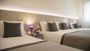 A hotel room with three beds, each with a pillow and a bedside table with a lamp and flowers.