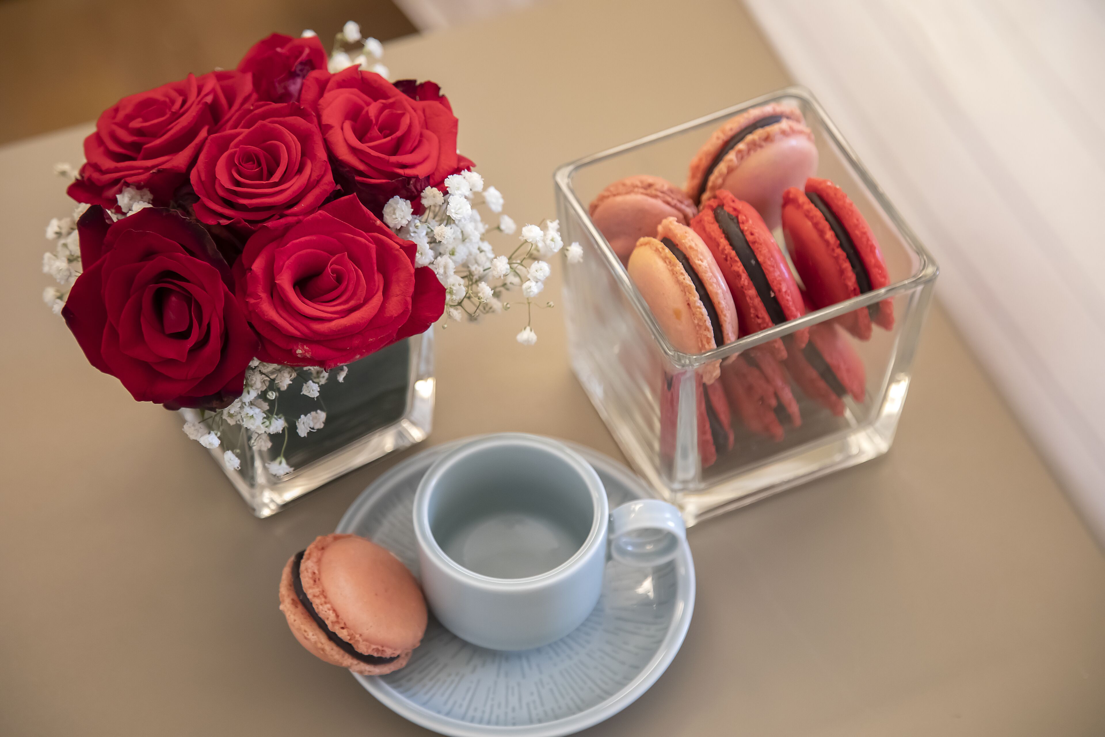 A table with a vase of red roses, a clear glass container with macarons, and a blue cup and saucer.