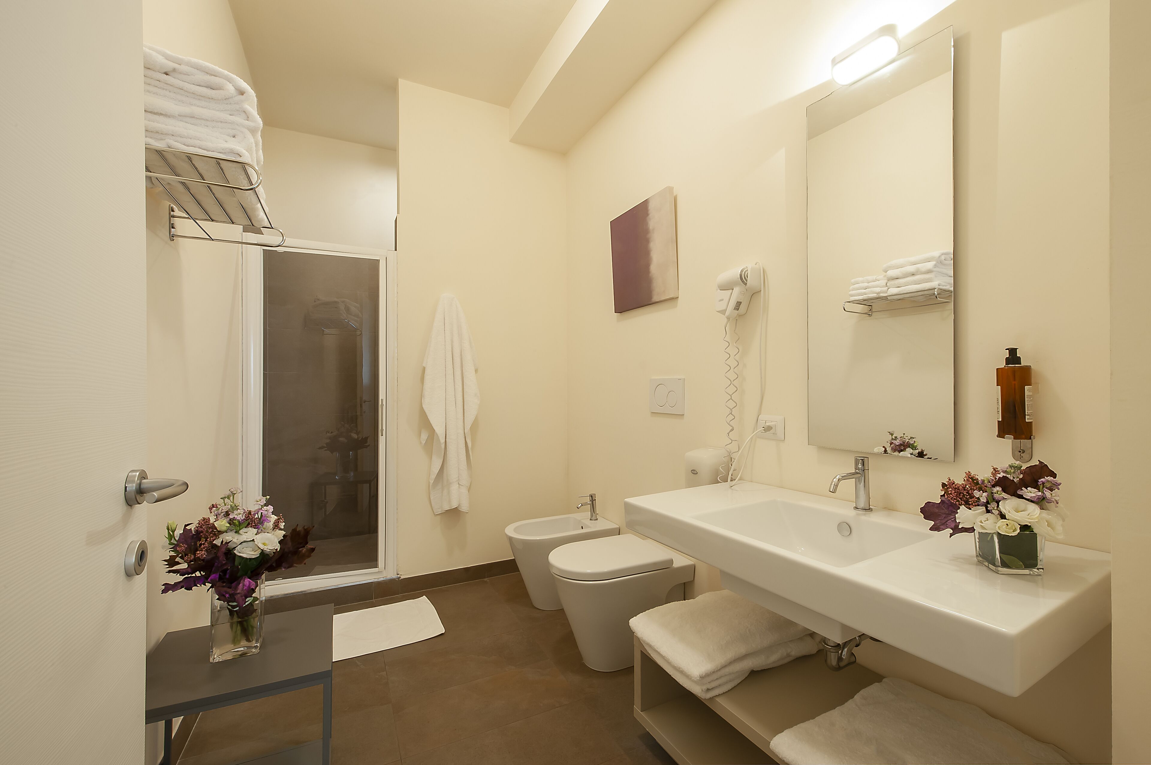A modern bathroom with a glass shower enclosure, a toilet, a bidet, a sink with a mirror, and a vase of flowers on the counter.