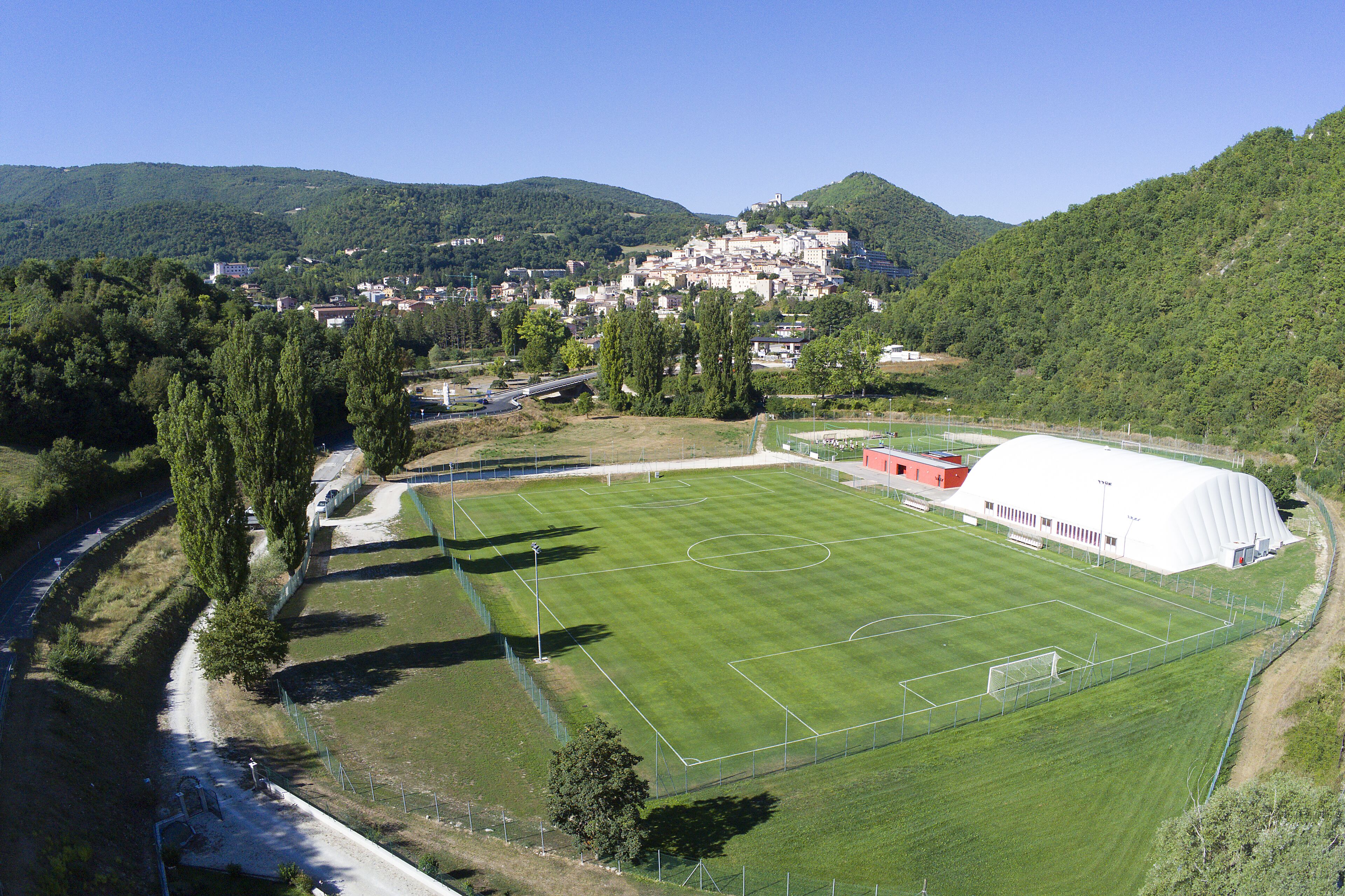 An aerial view of a soccer field with a large white building adjacent to it, surrounded by greenery and hills.