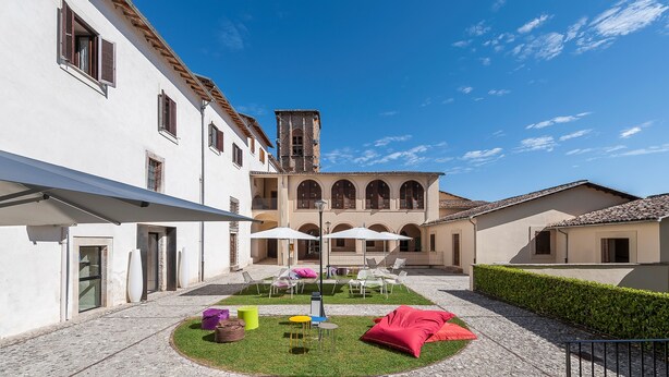 A courtyard with outdoor seating, a fountain, and a building with arched windows and a bell tower. - Hotel La Corte (Cascia)