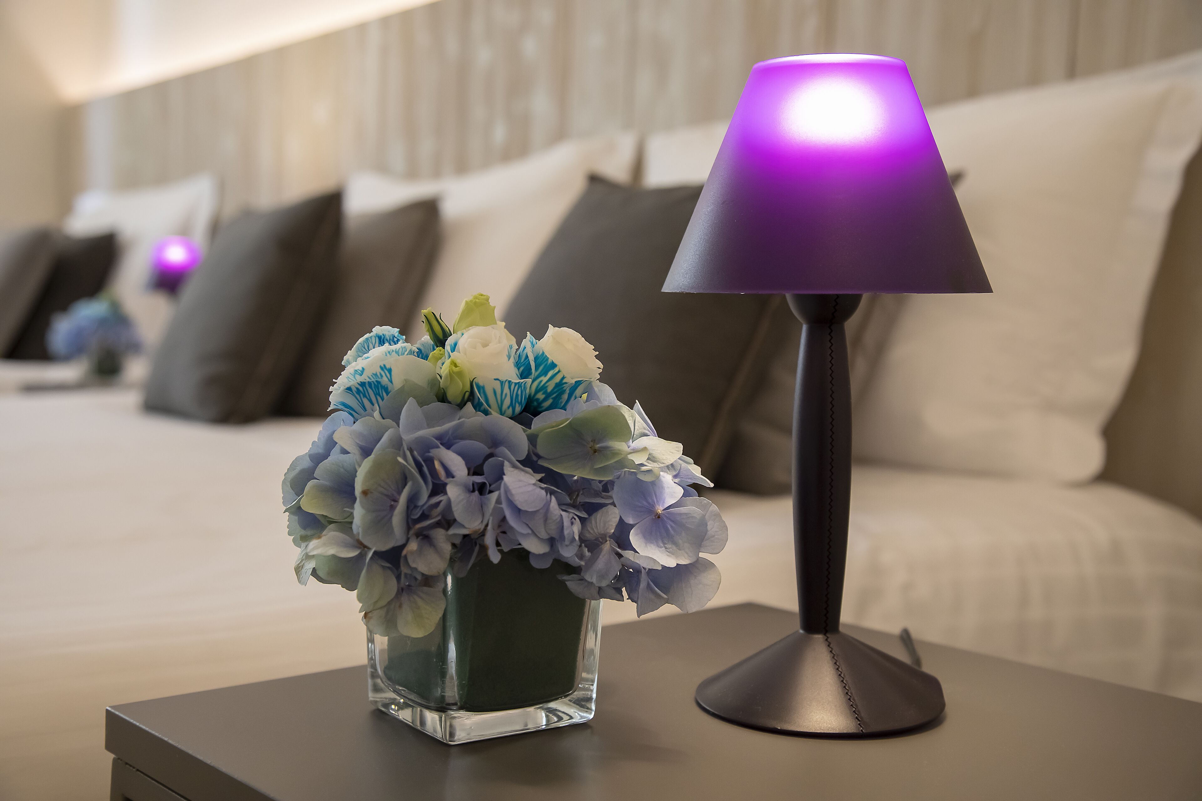 A modern bedroom with a bed, a bedside table with a lamp, and a vase of flowers.