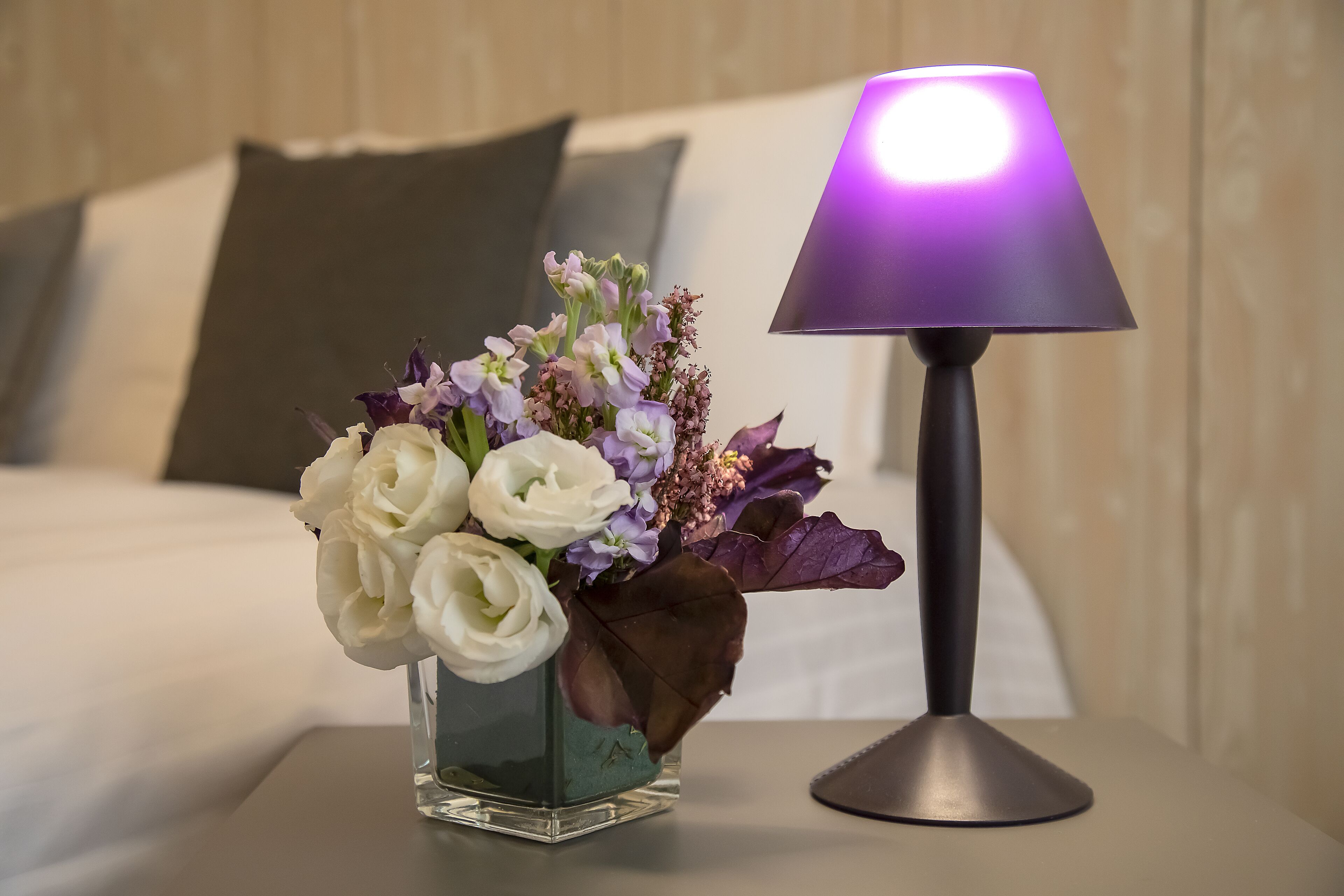A bed with a pillow, a vase with flowers, and a lamp on a bedside table.