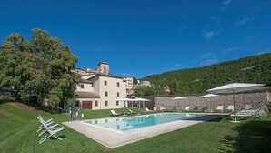 A pool area with lounge chairs, a building with a stone facade, and a hillside in the background. - Hotel La Corte (Cascia)
