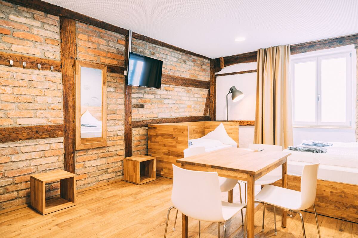 A room with a wooden table, white chairs, a TV mounted on a brick wall, and a bed with a window.