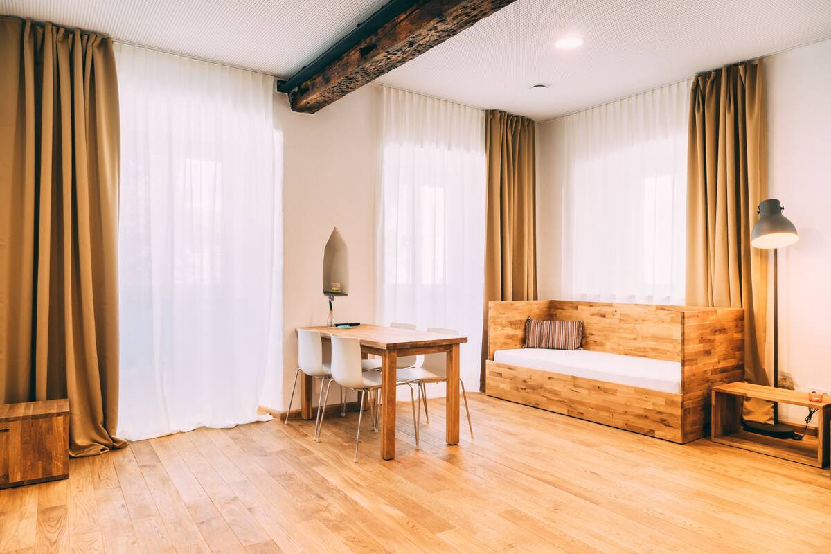 A room with a wooden floor, a wooden desk with chairs, a wooden bed, and a small table with a lamp.