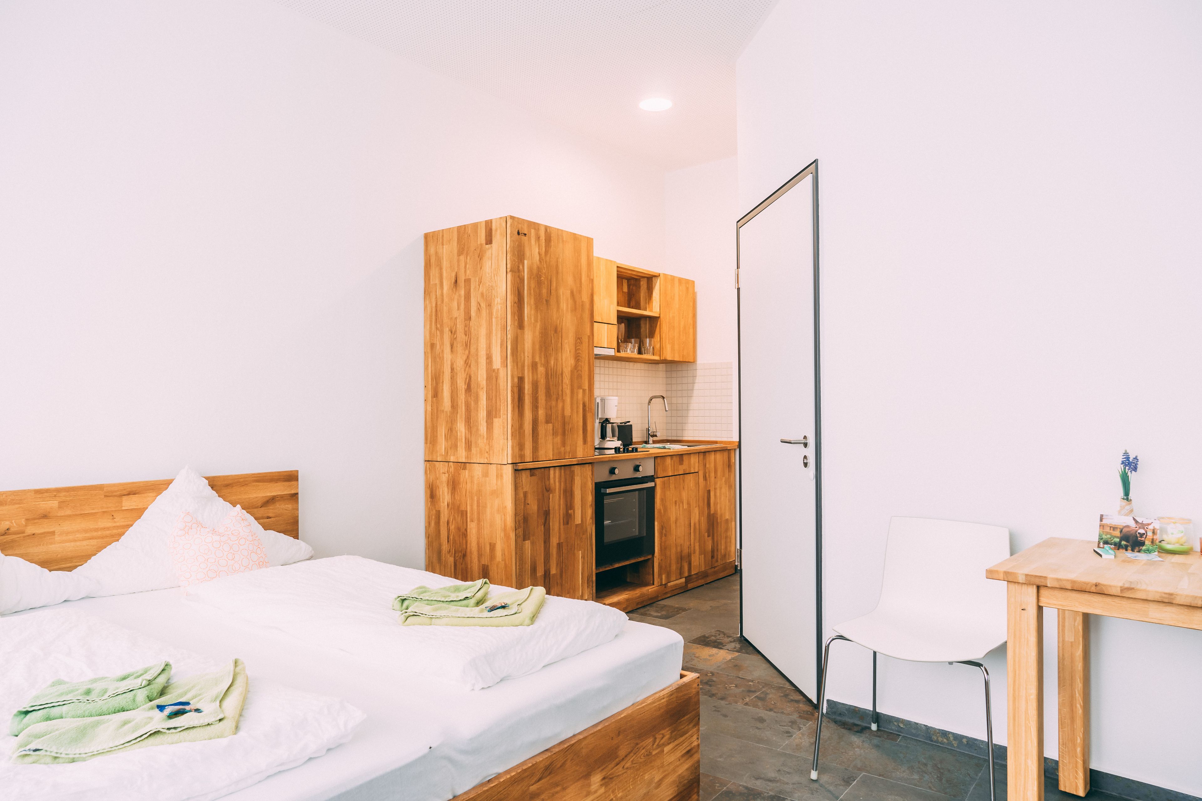 A bedroom with a wooden bed, a small kitchenette, and a white chair.