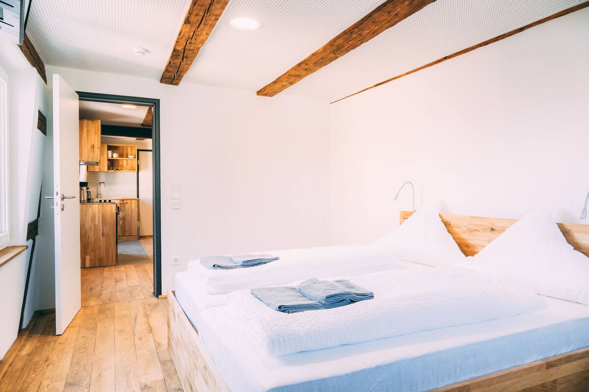 A bedroom with two beds, a wooden ceiling, and a kitchen in the background.