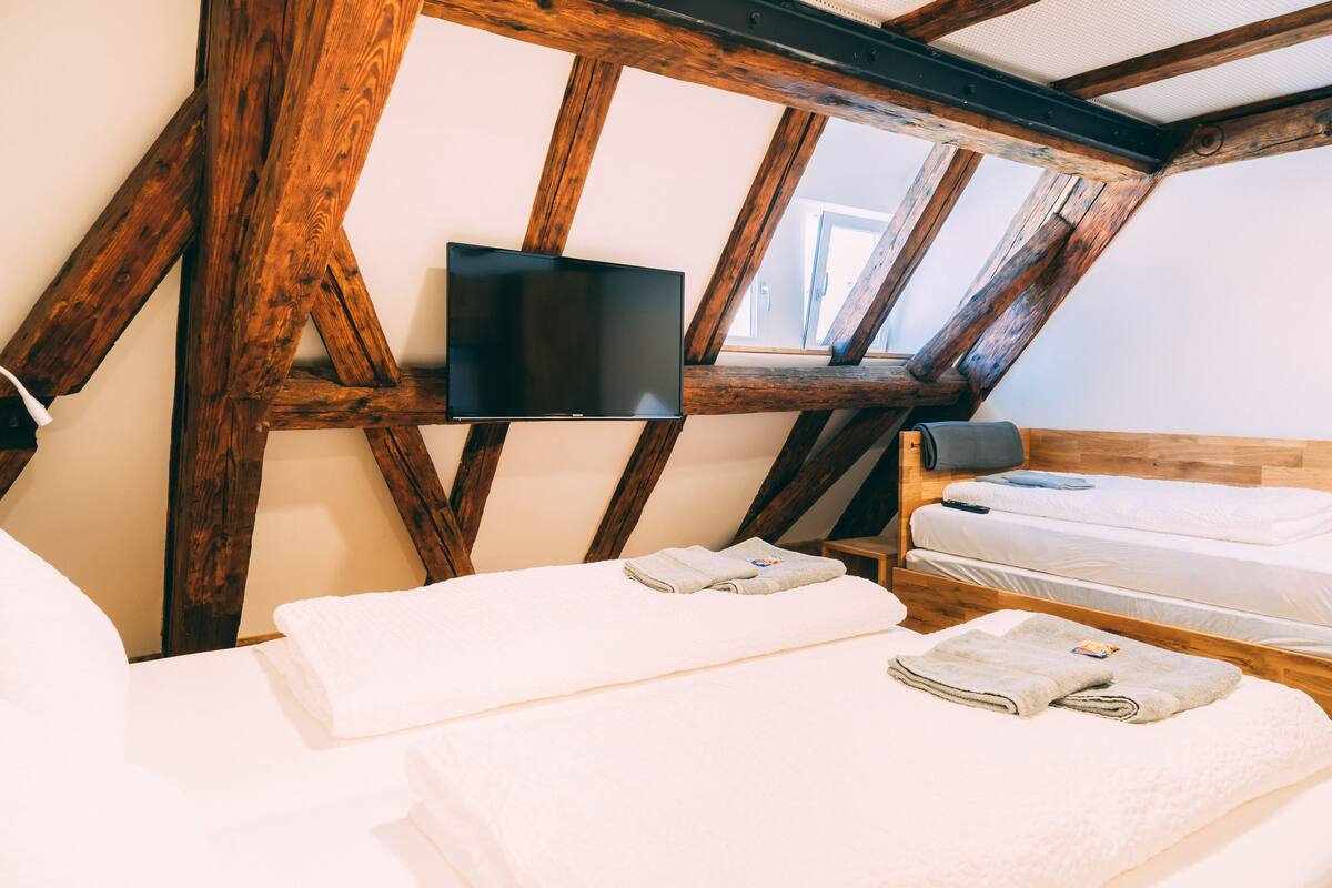 A room with a sloped ceiling, wooden beams, a flat-screen TV mounted on the wall, two beds with neatly folded towels, and a bedside table with a lamp.