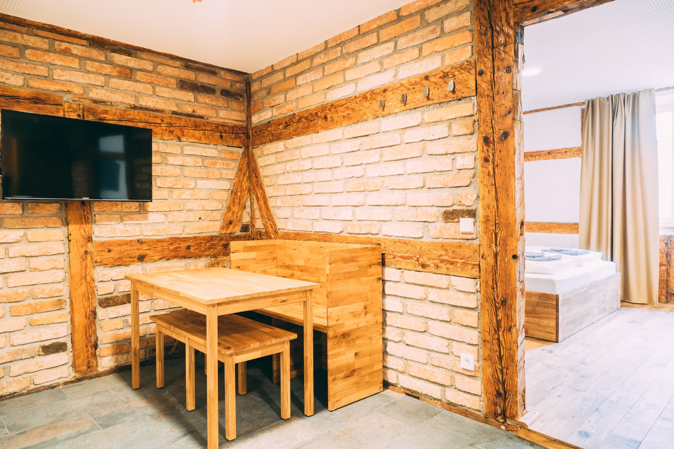 Una habitación con pared de ladrillo, una mesa de madera con sillas, un televisor de pantalla plana montado en la pared y una cama con colchón blanco y cortinas color beige.