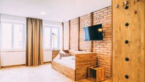 A modern bedroom with a wooden bed, a TV mounted on a brick wall, and a window with curtains. - Amtshaus Gut Hügle Boutique Apartments (Bad Waldsee)