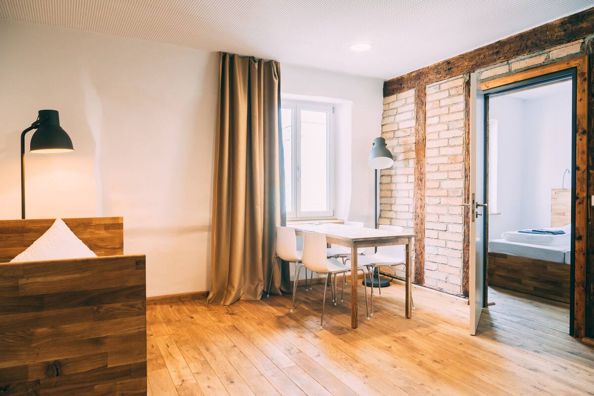 A modern room with a wooden floor, a white desk, two chairs, a black lamp, and a brick wall.