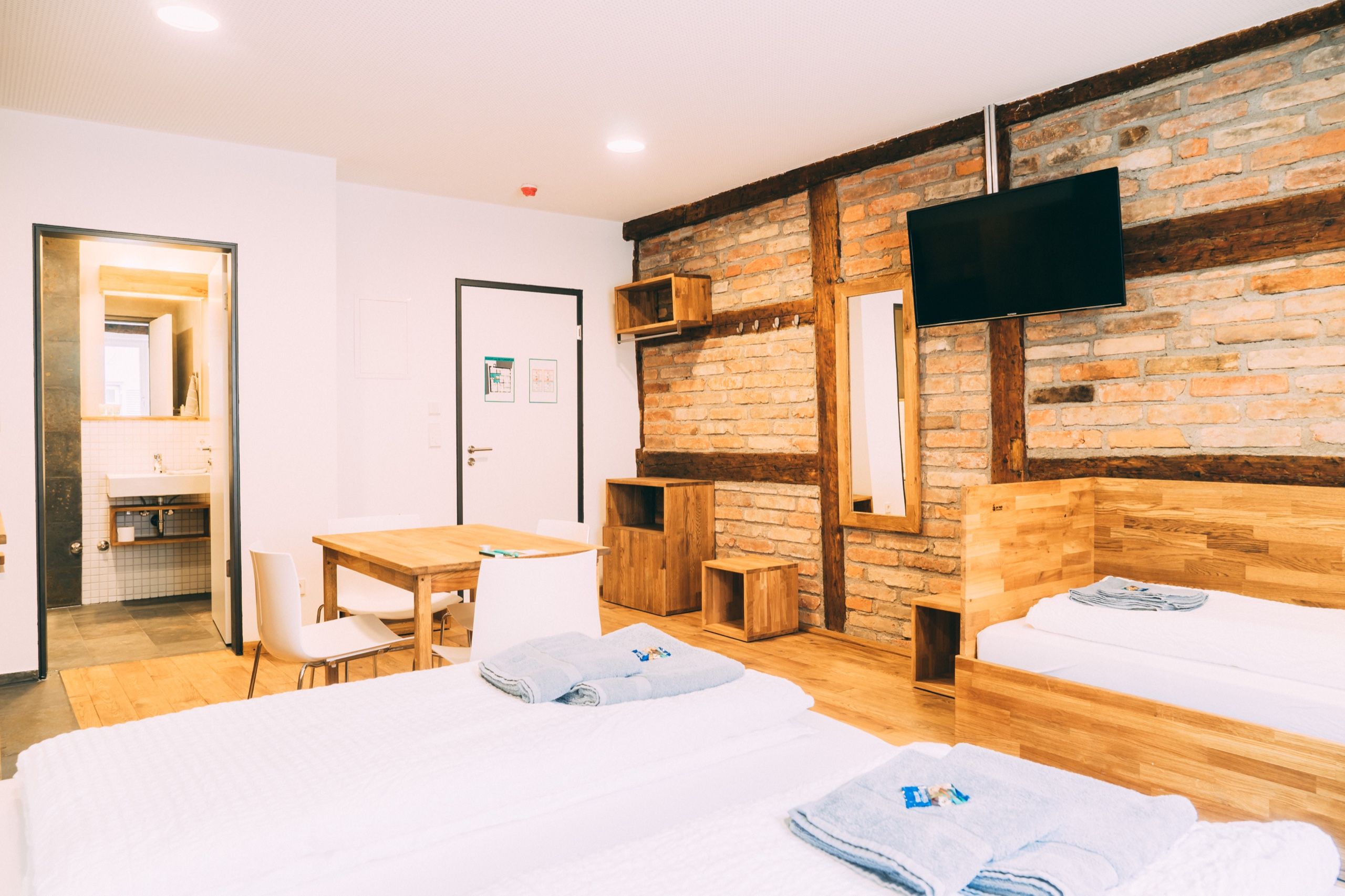 A hotel room with a wooden headboard, a flat-screen TV mounted on the brick wall, a wooden desk, and a white bed with towels.
