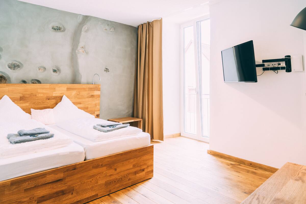 A modern hotel room with a wooden bed, a flat-screen TV mounted on the wall, and a large window with curtains.