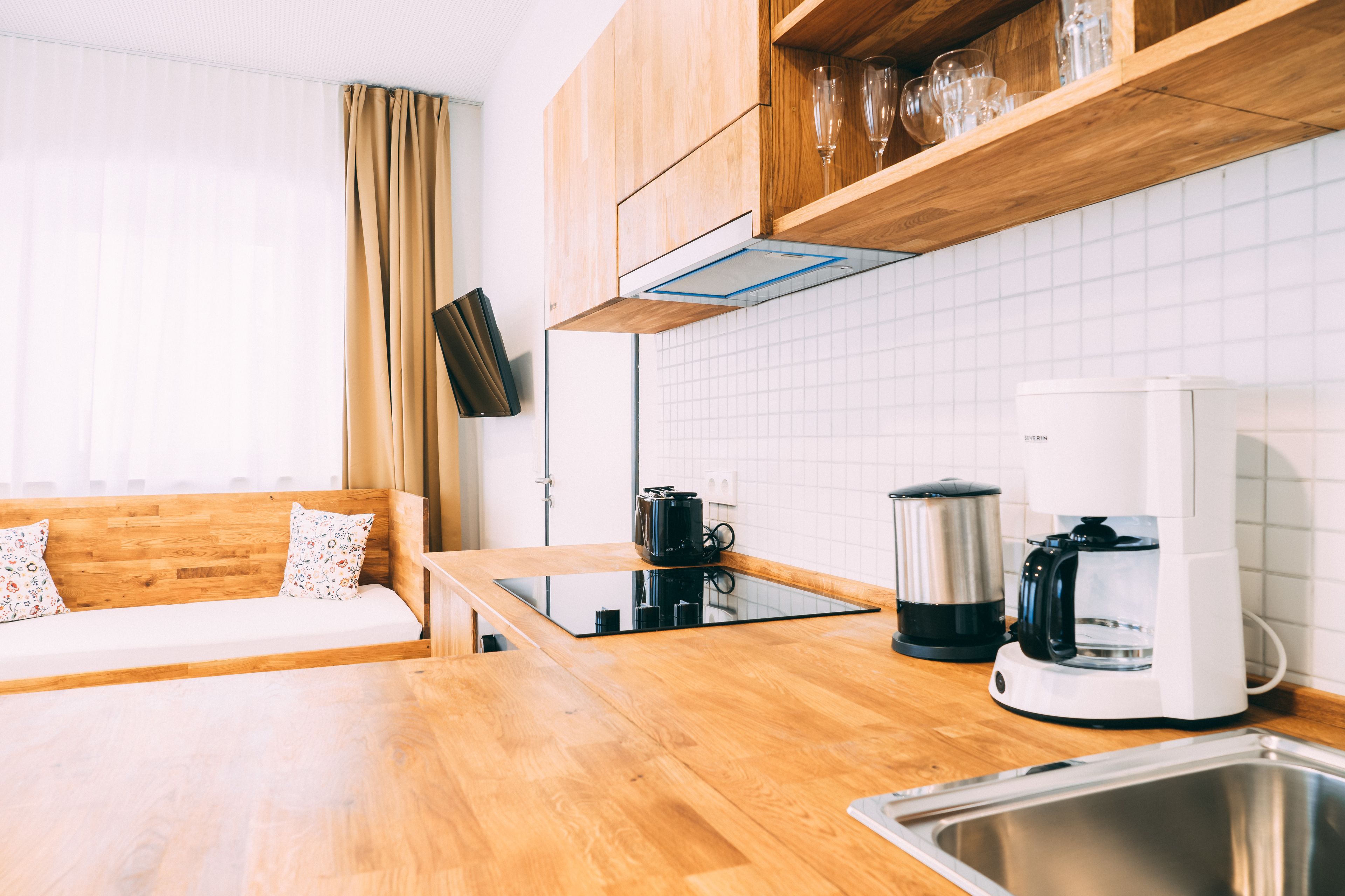 A modern kitchen with wooden cabinets, a built-in stove, a coffee maker, and a window with curtains.