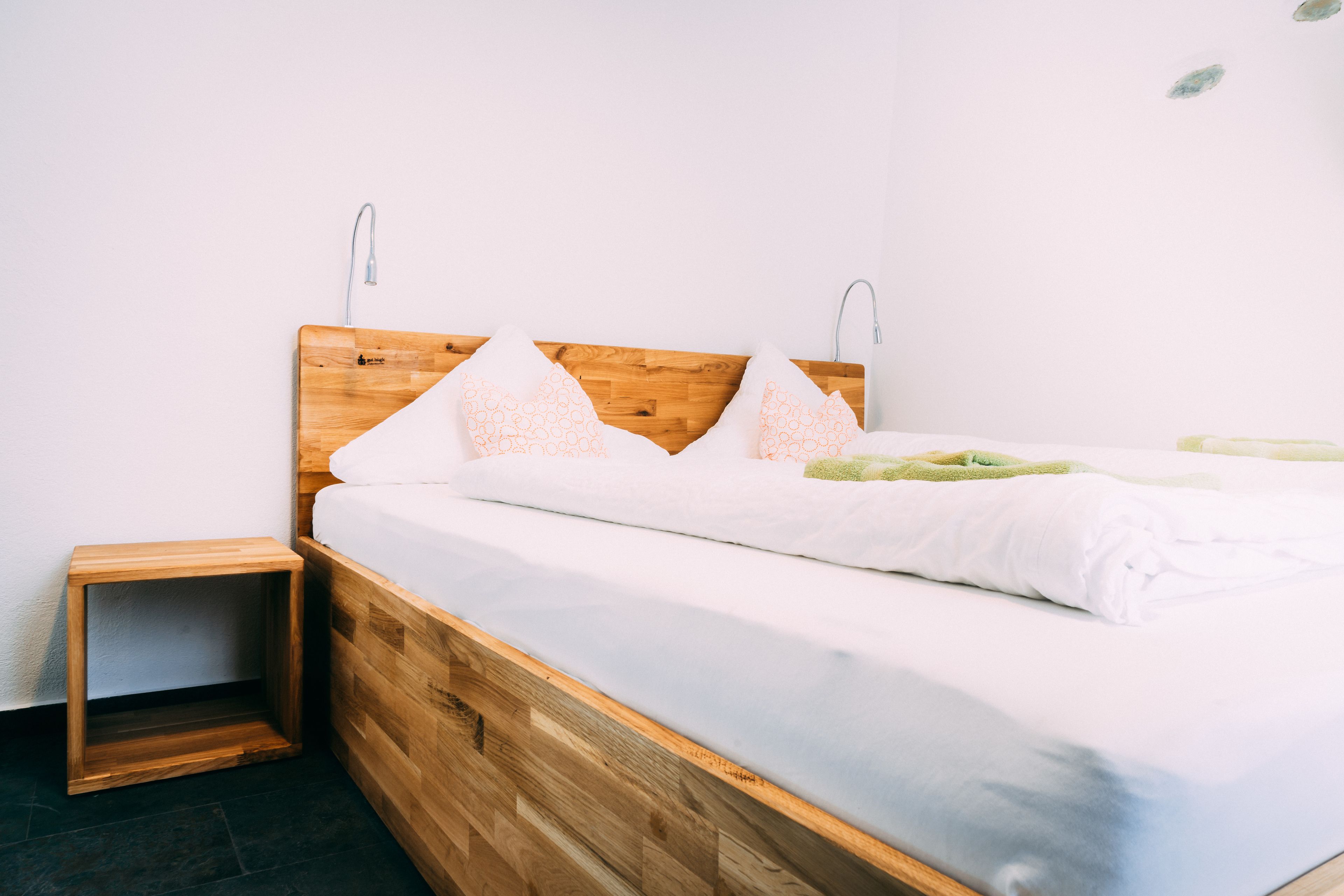 Un lit bien fait, avec une tête de lit en bois, une petite table de chevet en bois et un mur blanc sobre.