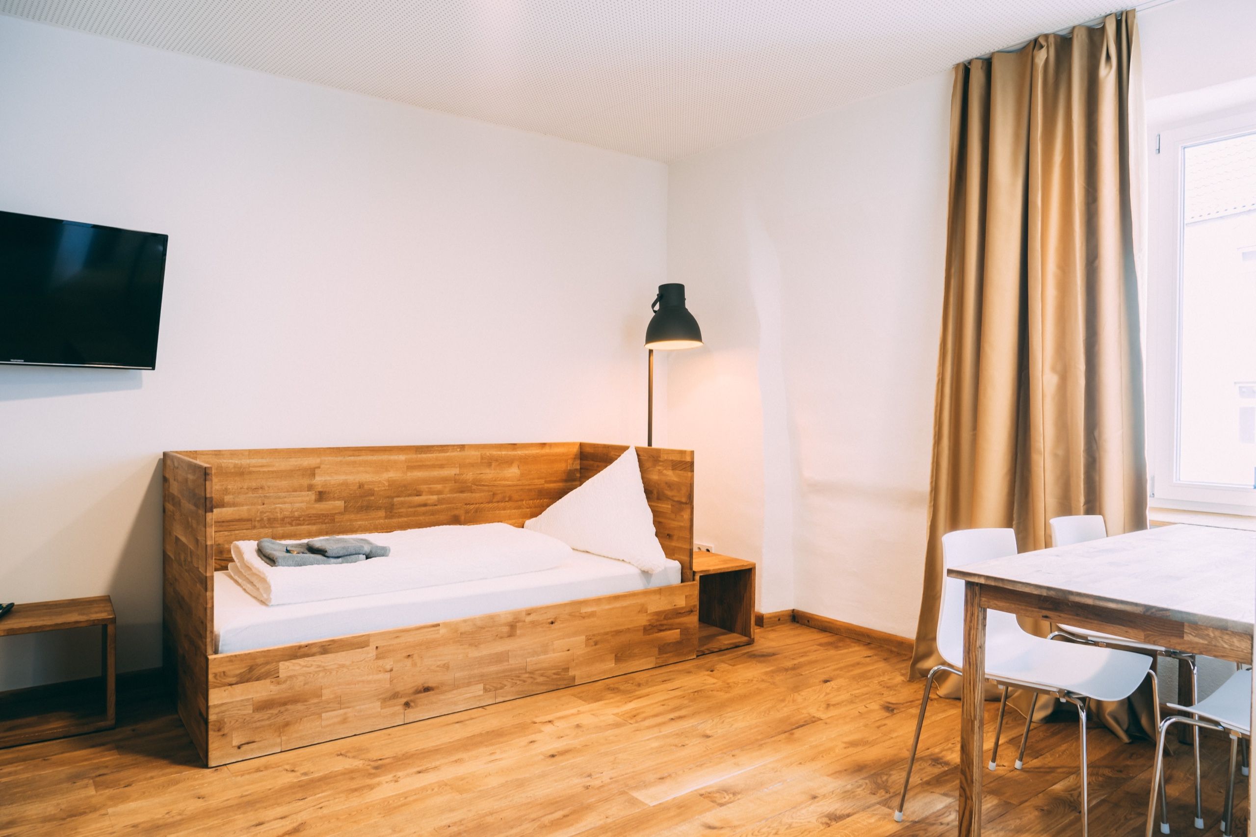 Una habitación con una cama de madera, un escritorio, una silla, una lámpara y un televisor en la pared.