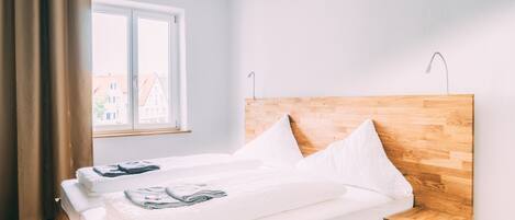 A hotel room with a wooden headboard, a large window with curtains, a bed with white linens, and a small wooden nightstand.