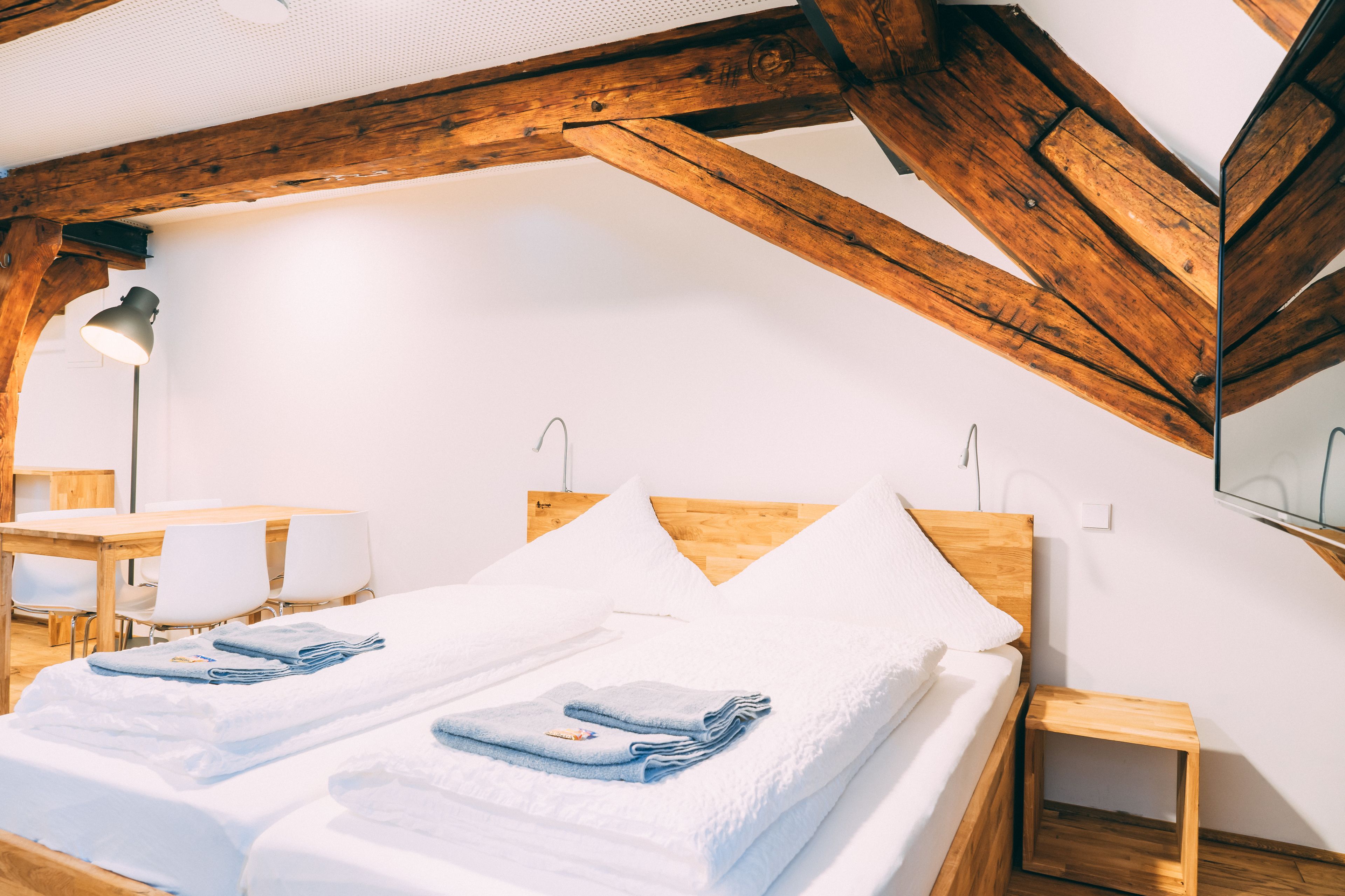 Una habitación con techo de madera, una cama con ropa blanca, una pequeña mesa de madera y una silla.