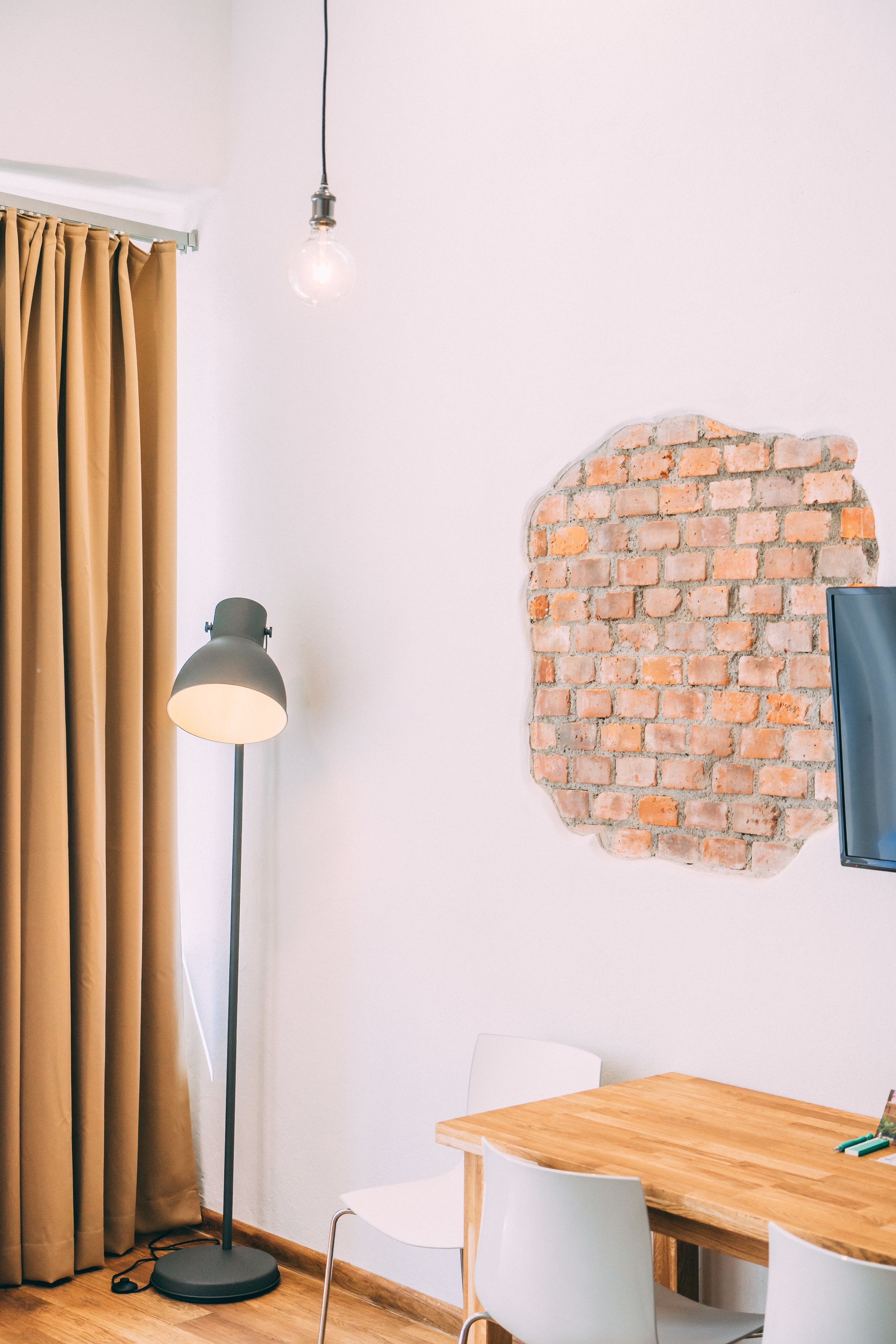 A room with a brick wall, a wooden table, white chairs, a floor lamp, and a curtain.