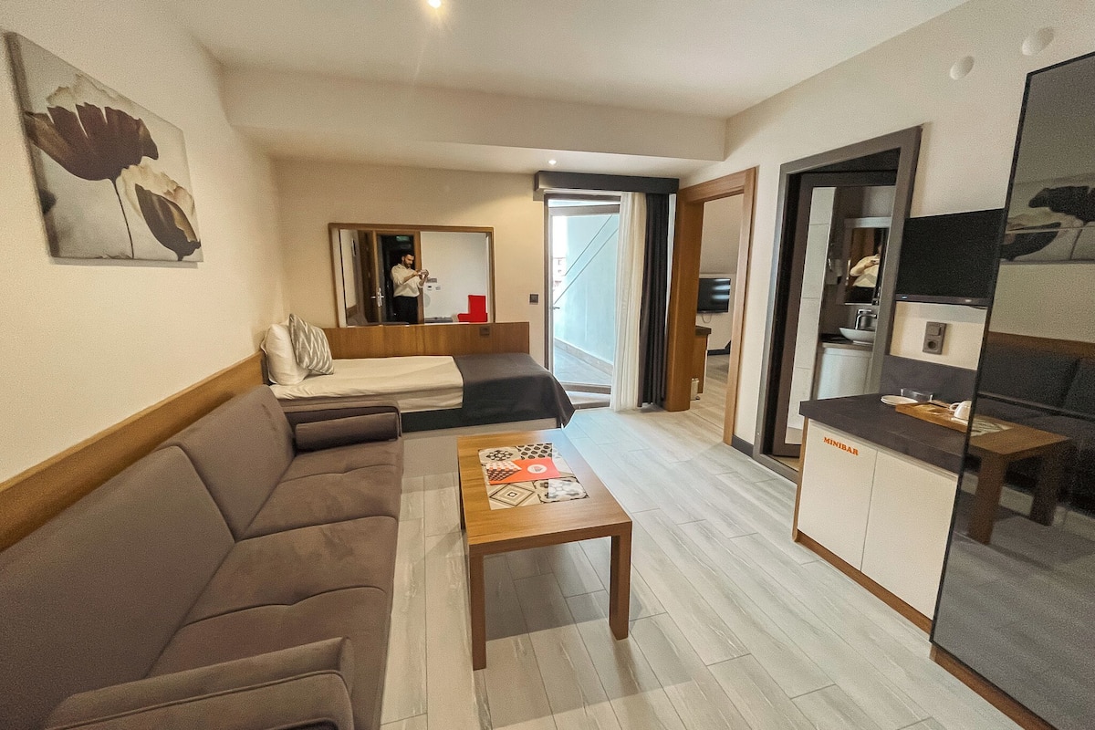 A modern hotel room with a sofa, a wooden coffee table, a bed, a kitchenette, and a large mirror.