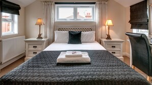 A neatly made bed with a dark patterned bedspread, two bedside tables with lamps, a window with curtains, and a view of buildings outside. - Arlana Guest House (Cleethorpes)