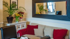 A living room with a beige sofa, red cushions, a black-framed mirror, and a potted plant with yellow flowers. - Arlana Guest House (Cleethorpes)