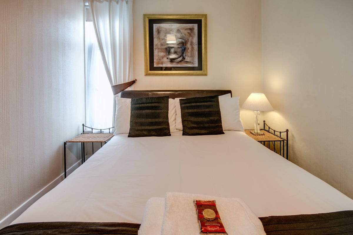 A hotel room with a large bed, a framed artwork on the wall, a bedside table with a lamp, and a chair by the window.