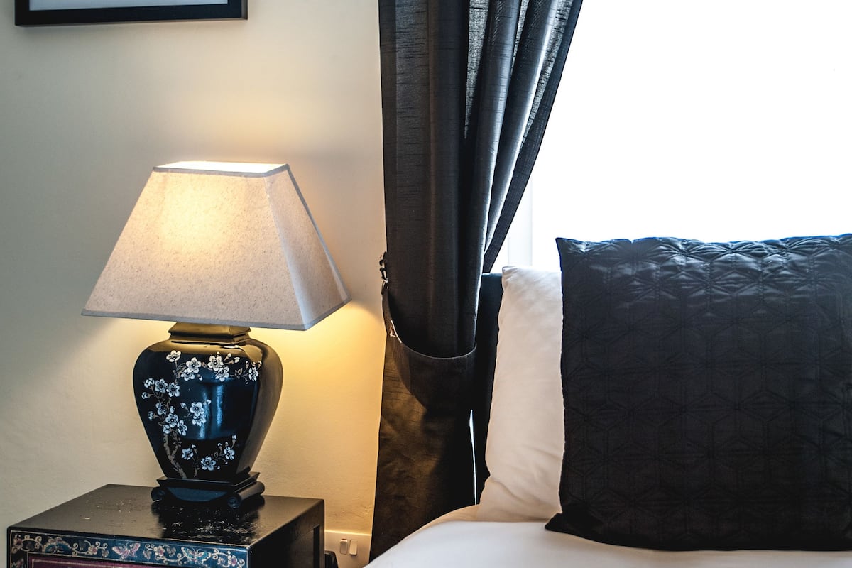 A neatly made bed with a white comforter and a dark pillow, a bedside table with a lamp, and a framed picture on the wall.