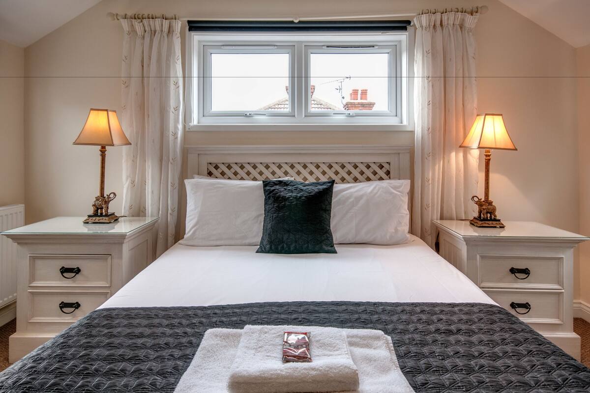 A neatly arranged bedroom with a large bed, two bedside tables with lamps, a window with curtains, and a view of buildings outside.