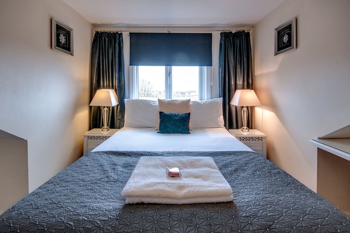 A hotel room with a large bed, two bedside lamps, a window with curtains, and a small table with a decorative item.