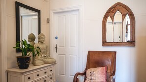 A hallway with a white door, a wooden chair with a cushion, a potted plant, a white dresser with drawers, and a mirror with a wooden frame. - Arlana Guest House (Cleethorpes)