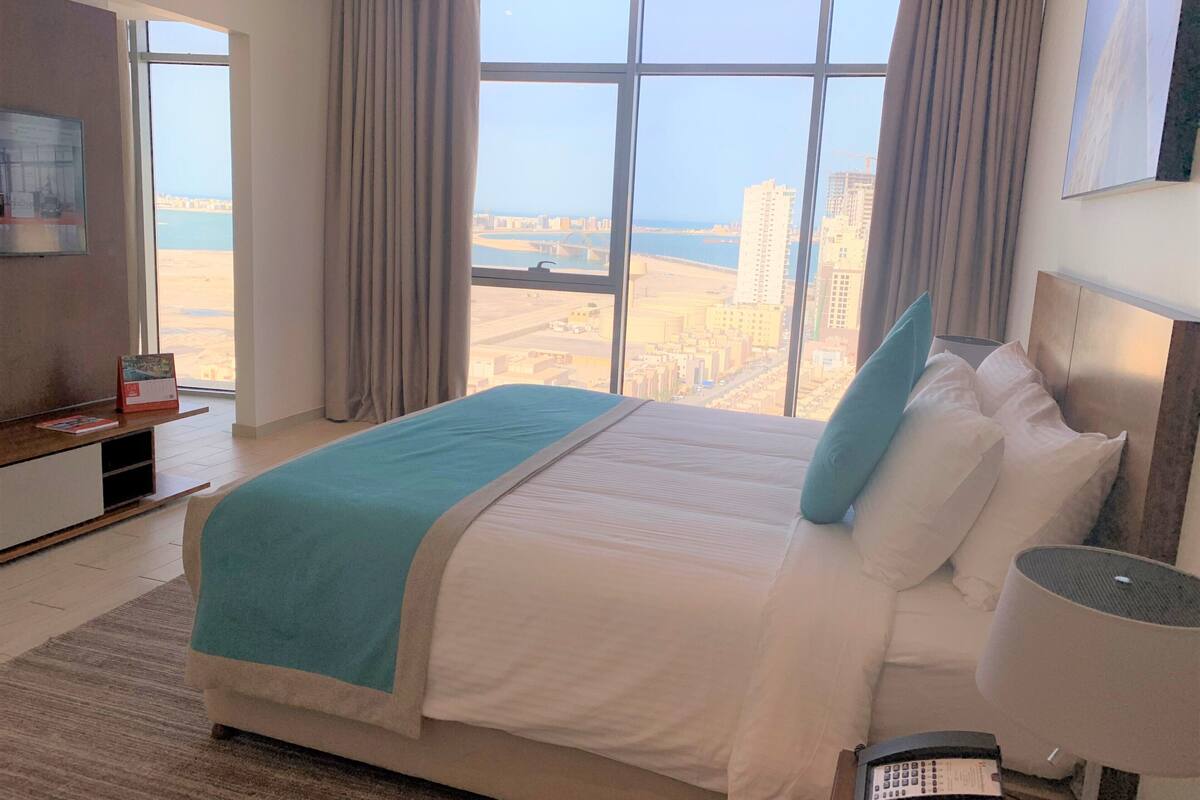 A hotel room with a large bed, a desk with a computer, a TV, and a view of the city and water.