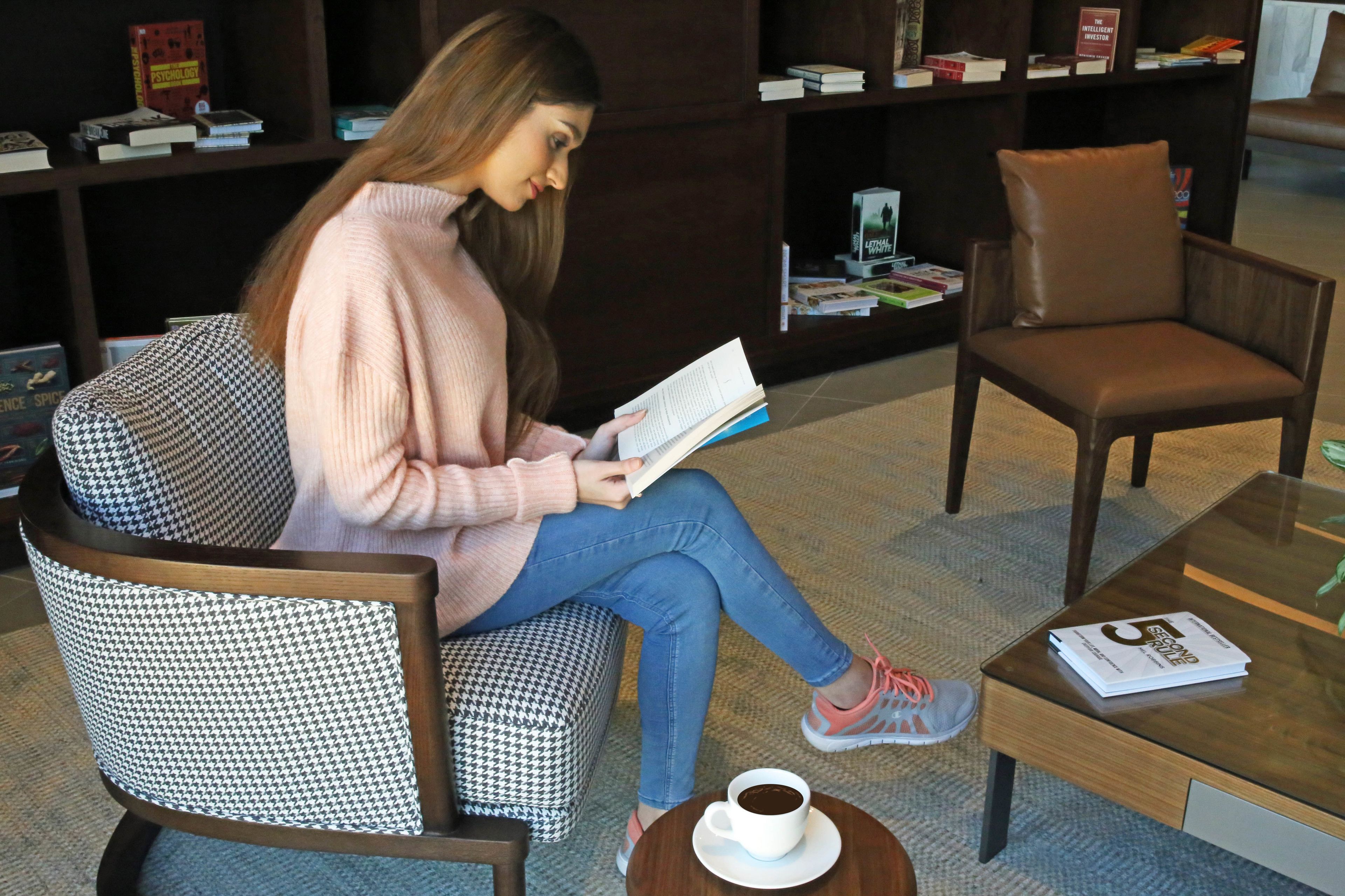 Eine Person sitzt auf einem Stuhl mit Hunde-Zahn-Muster und liest ein Buch, mit einer Kaffeetasse auf einem kleinen Tisch in der Nähe.