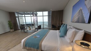 Ein modernes Hotelzimmer mit einem großen Bett, einem Schreibtisch, einem Sessel, einer Zimmerpflanze und Meerblick durch große Fenster.