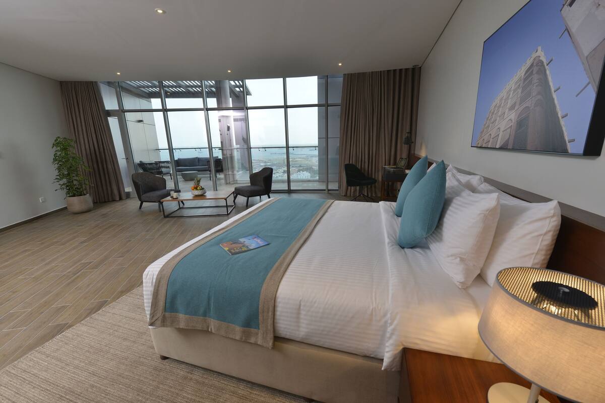 A modern hotel room with a large bed, a desk, a chair, a potted plant, and a view of the ocean through large windows.
