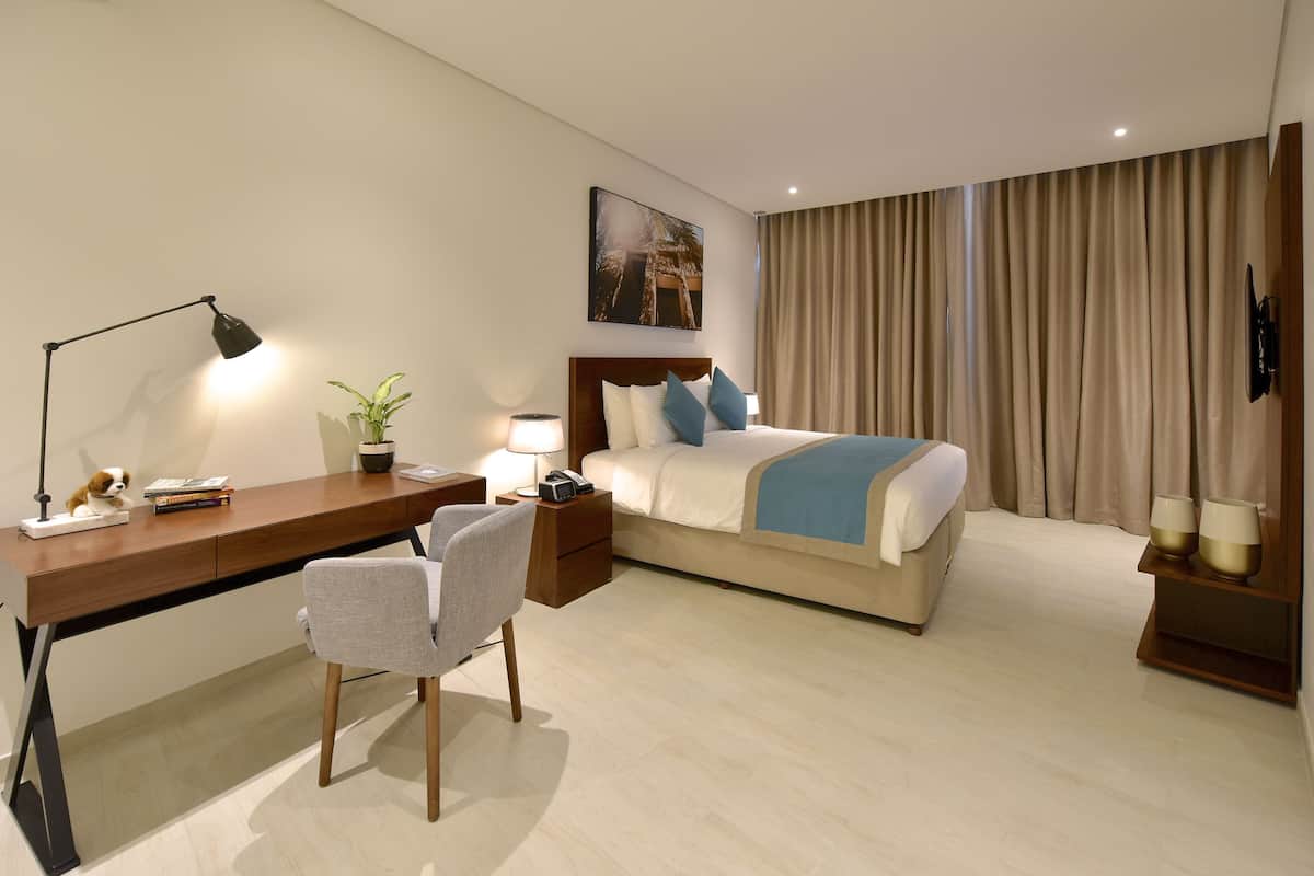 A hotel room with a wooden desk, a chair, a bed with a blue blanket, a nightstand with a lamp, a potted plant, and a painting on the wall.