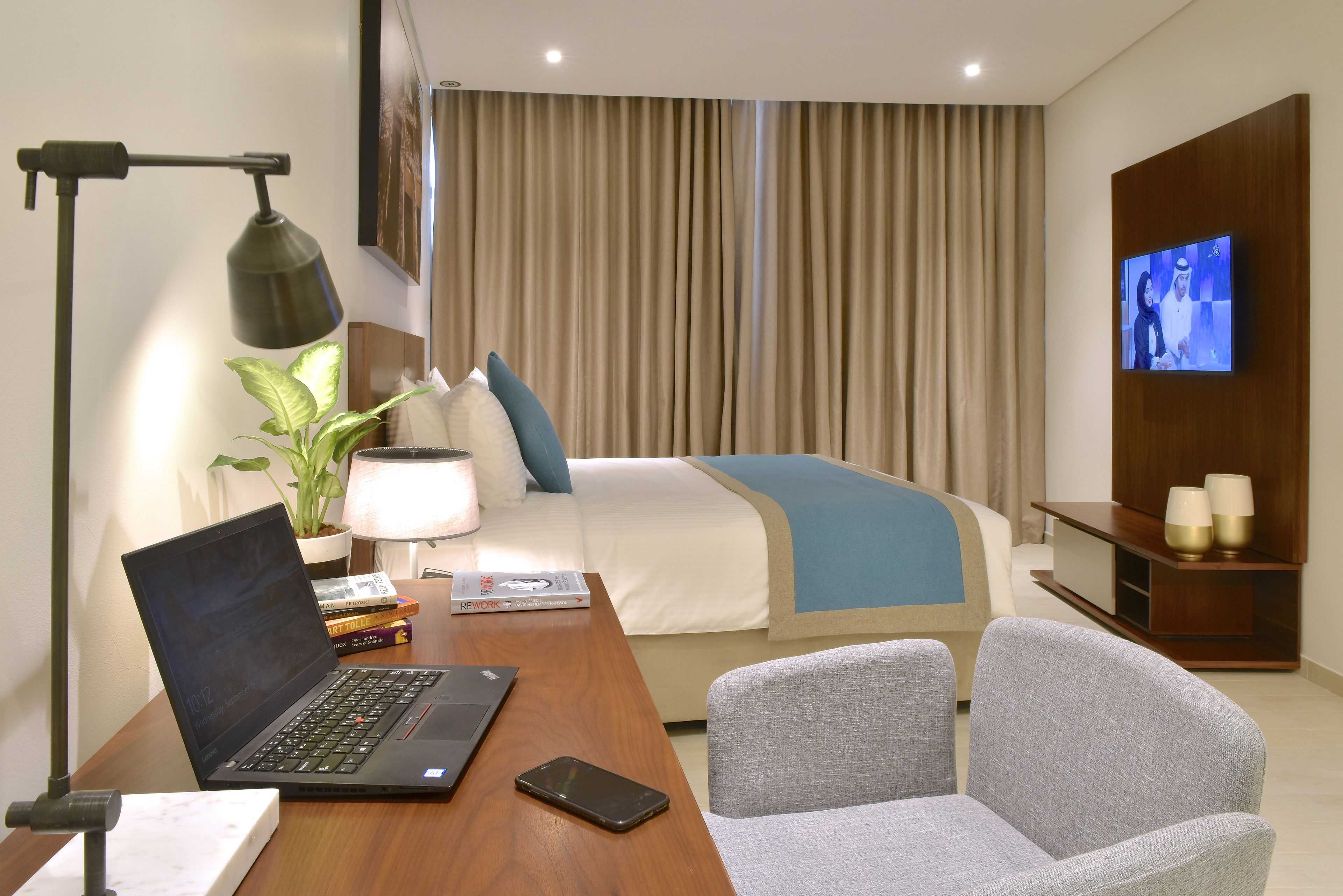 A hotel room with a bed, a desk with a laptop, a chair, a TV, and a lamp.